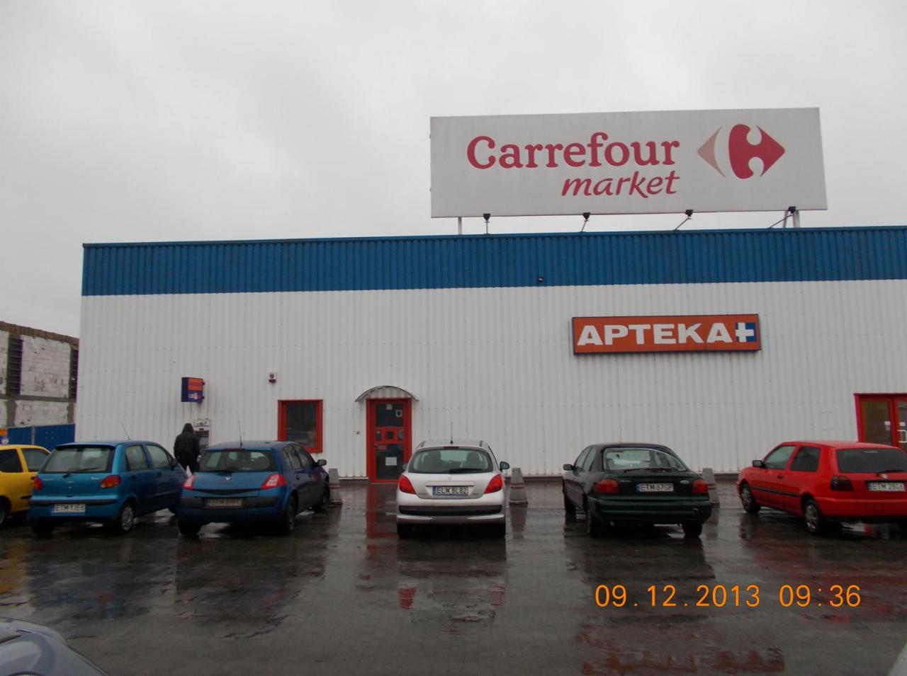 Fasada budynku Carrefour Market z apteką, widok z parkingu w deszczowy dzień, z zaparkowanymi samochodami i datą 09.12.2013 09:36 w prawym dolnym rogu.