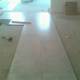 Układanie jasnych, kwadratowych płytek podłogowych w korytarzu, z poziomnicą i gumowym młotkiem widocznymi na surowym podłożu.