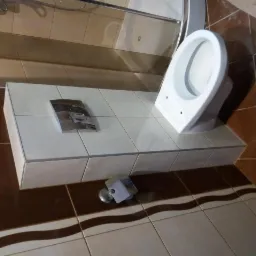 Podwieszana toaleta z chromowanym przyciskiem spłukującym, zamontowana na podwyższeniu z płytek, widoczny uchwyt na papier toaletowy i fragment kabiny prysznicowej.