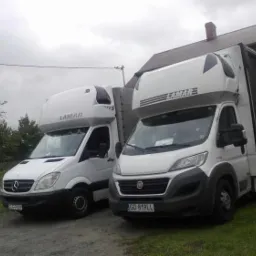 Dwa białe busy dostawcze marki Mercedes i Fiat z nadbudówką i logo 'LAMAR' zaparkowane obok zielonego budynku w pochmurny dzień.