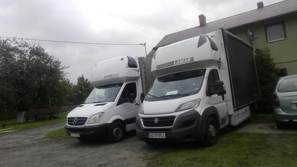 Dwa białe busy dostawcze marki Mercedes i Fiat z nadbudówką i logo 'LAMAR' zaparkowane obok zielonego budynku w pochmurny dzień.