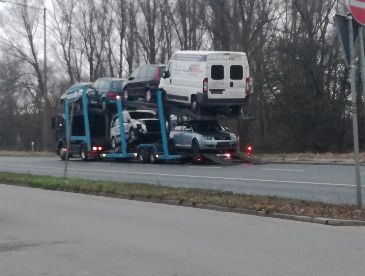 Laweta transportuje kilka samochodów i busa na autostradzie w pochmurny dzień. Widoczny napis 'HEIMA' na busie.