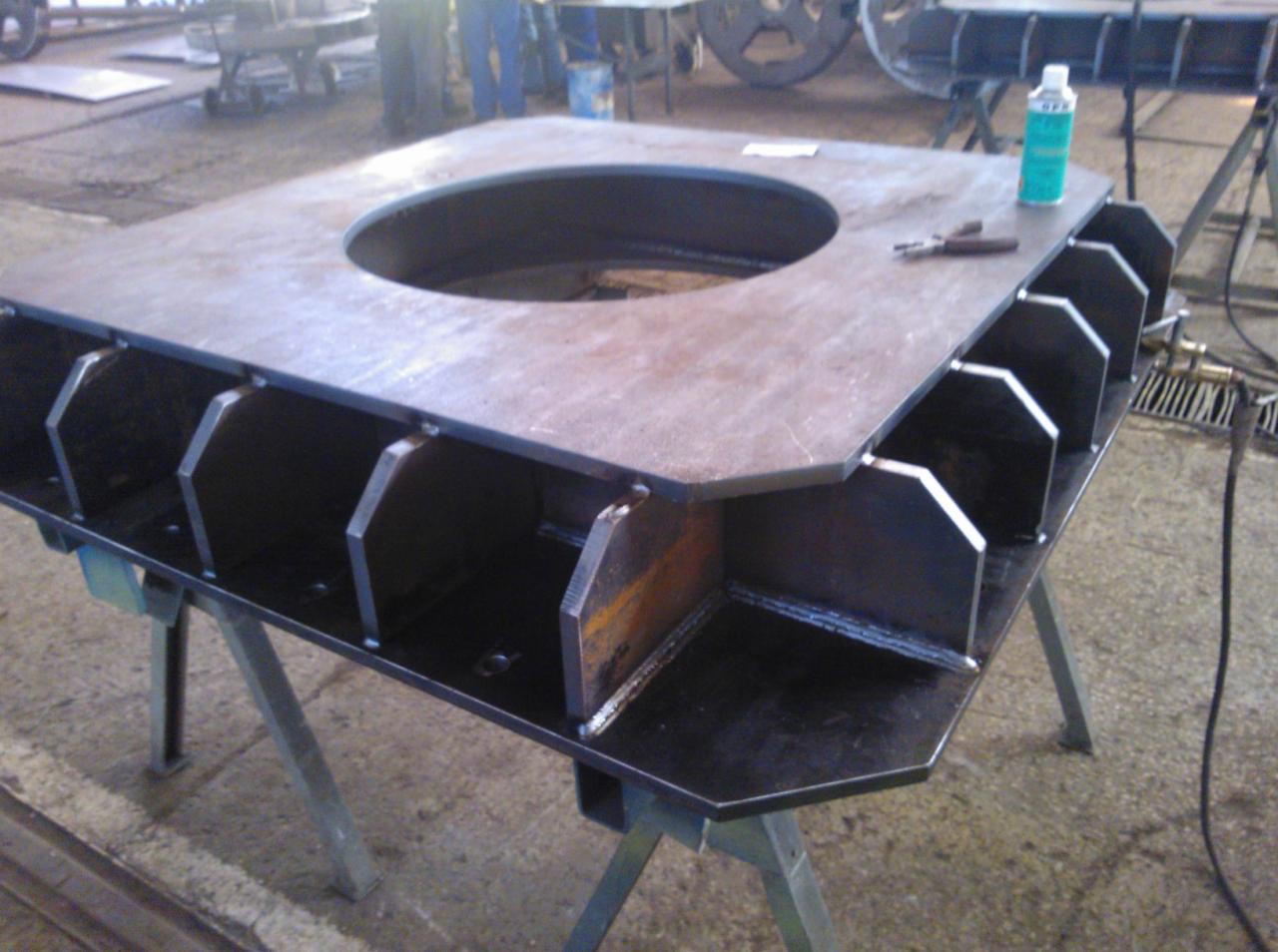 Konstrukcja metalowa spawana z otworem centralnym i wzmocnieniami, umieszczona na metalowym stojaku w hali produkcyjnej, obok leży butelka z preparatem.