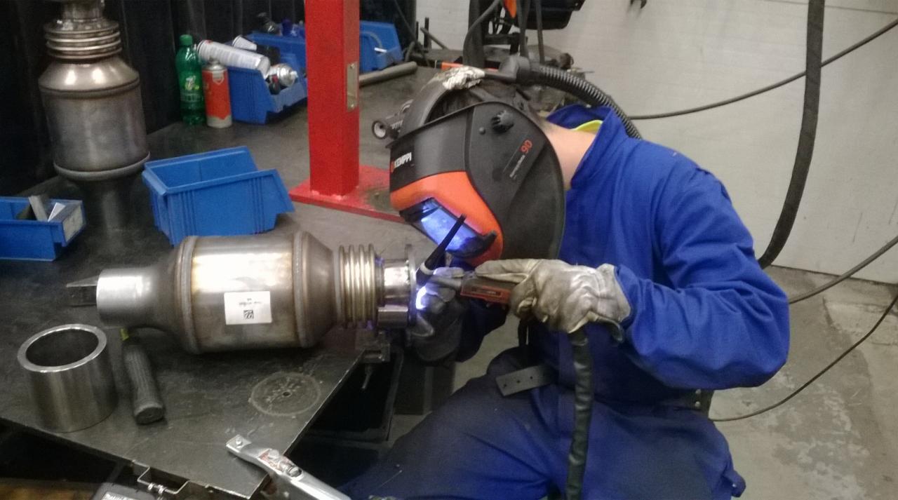 Pracownik w niebieskim kombinezonie i masce spawalniczej łączy metalowe elementy za pomocą spawarki, widoczne iskry i płomień.