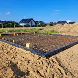 Zbrojenie płyty fundamentowej na piasku, zabezpieczone folią, z wystającymi rurami kanalizacyjnymi i żółtymi osłonami, w tle domy jednorodzinne i hałda ziemi.