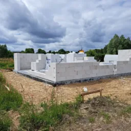 Wznoszenie ścian parterowego budynku z białych bloczków, widoczne fundamenty i częściowo rozpakowane palety z materiałem budowlanym na działce w trakcie budowy.