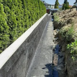 Świeżo zbudowany, długi mur oporowy z betonu komórkowego, zabezpieczony czarną izolacją bitumiczną, biegnący wzdłuż wysokich, zielonych tuj i ziemnego wykopu.