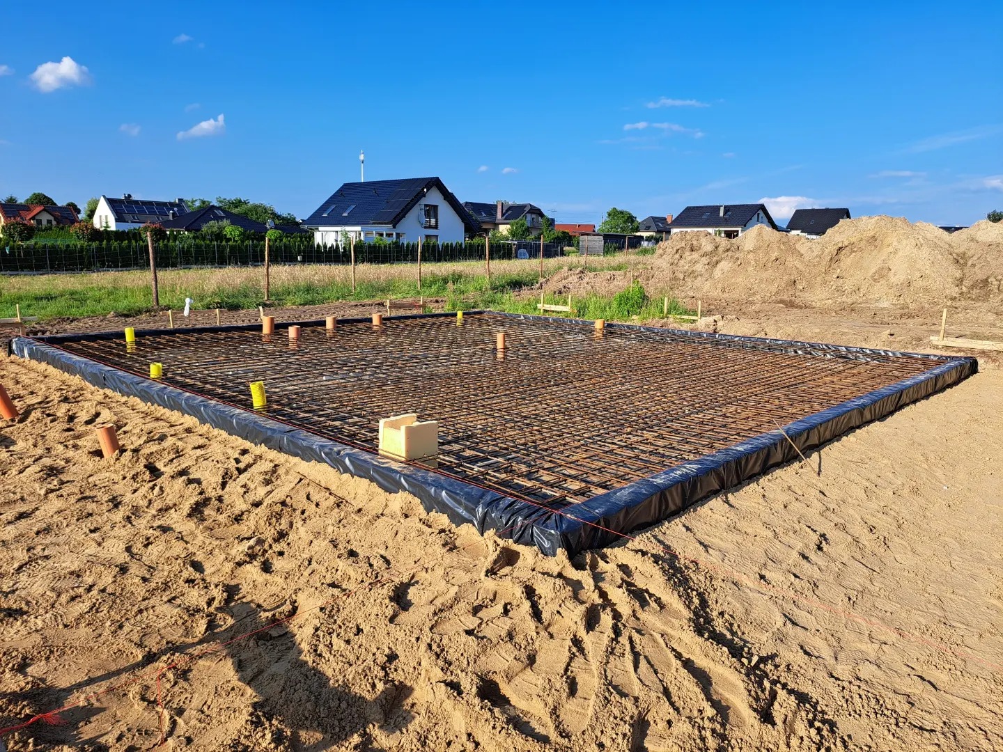 Zbrojenie płyty fundamentowej na piasku, zabezpieczone folią, z wystającymi rurami kanalizacyjnymi i żółtymi osłonami, w tle domy jednorodzinne i hałda ziemi.