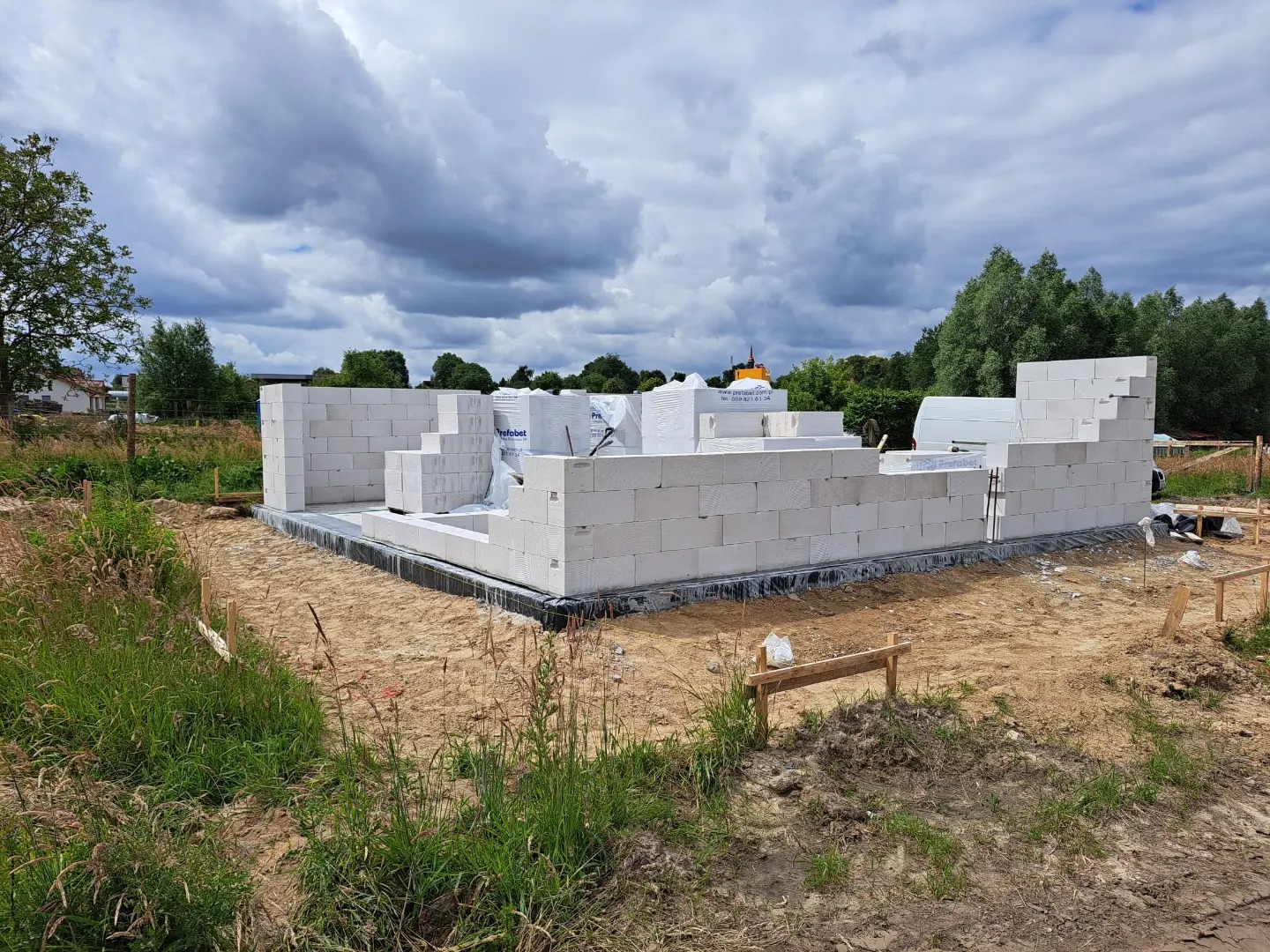 Wznoszenie ścian parterowego budynku z białych bloczków, widoczne fundamenty i częściowo rozpakowane palety z materiałem budowlanym na działce w trakcie budowy.