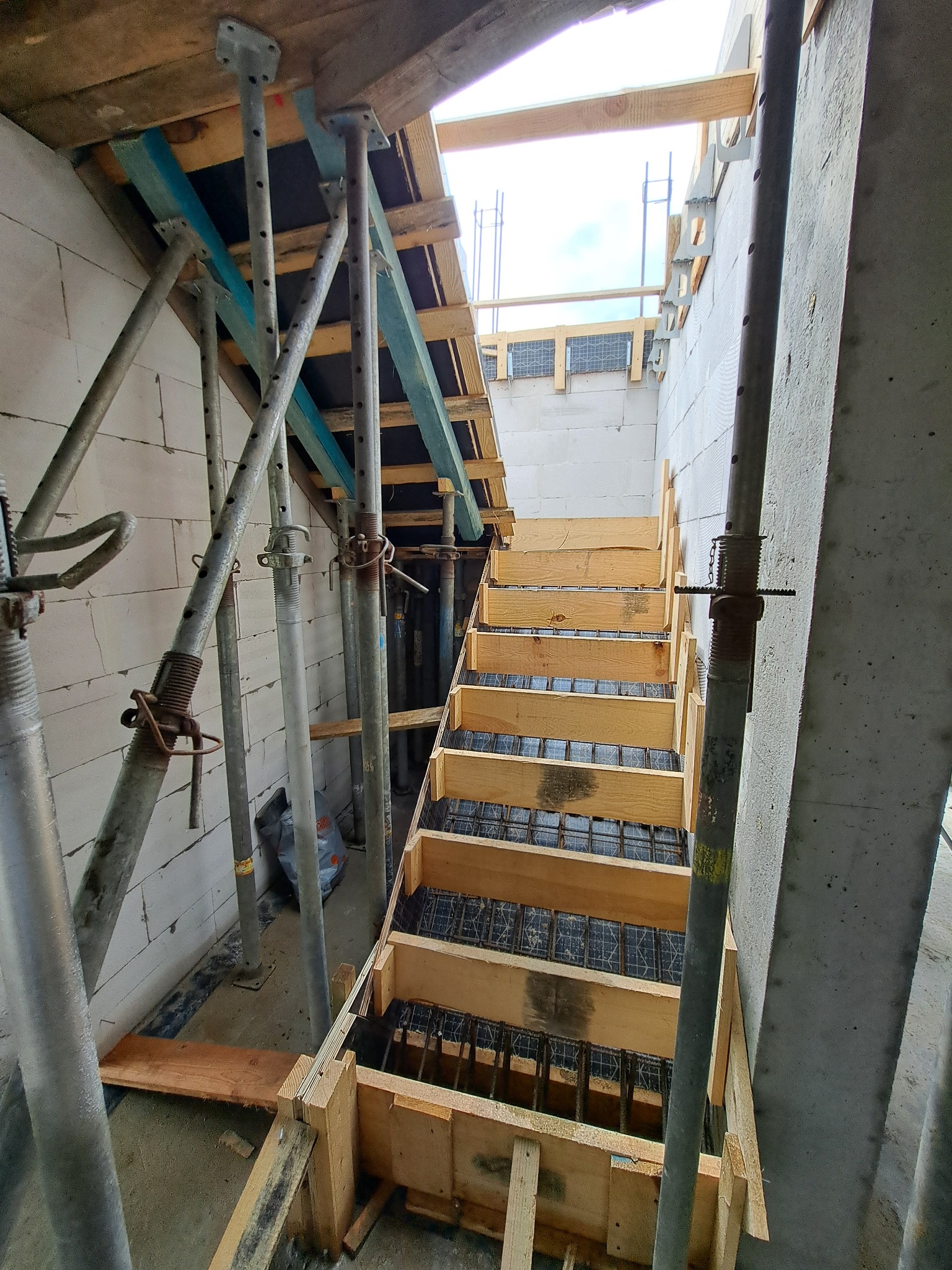 Wnętrze budynku w trakcie budowy, widoczne betonowe schody w drewnianym szalunku, wsparte na metalowych stemplach, ściany z bloczków betonowych.