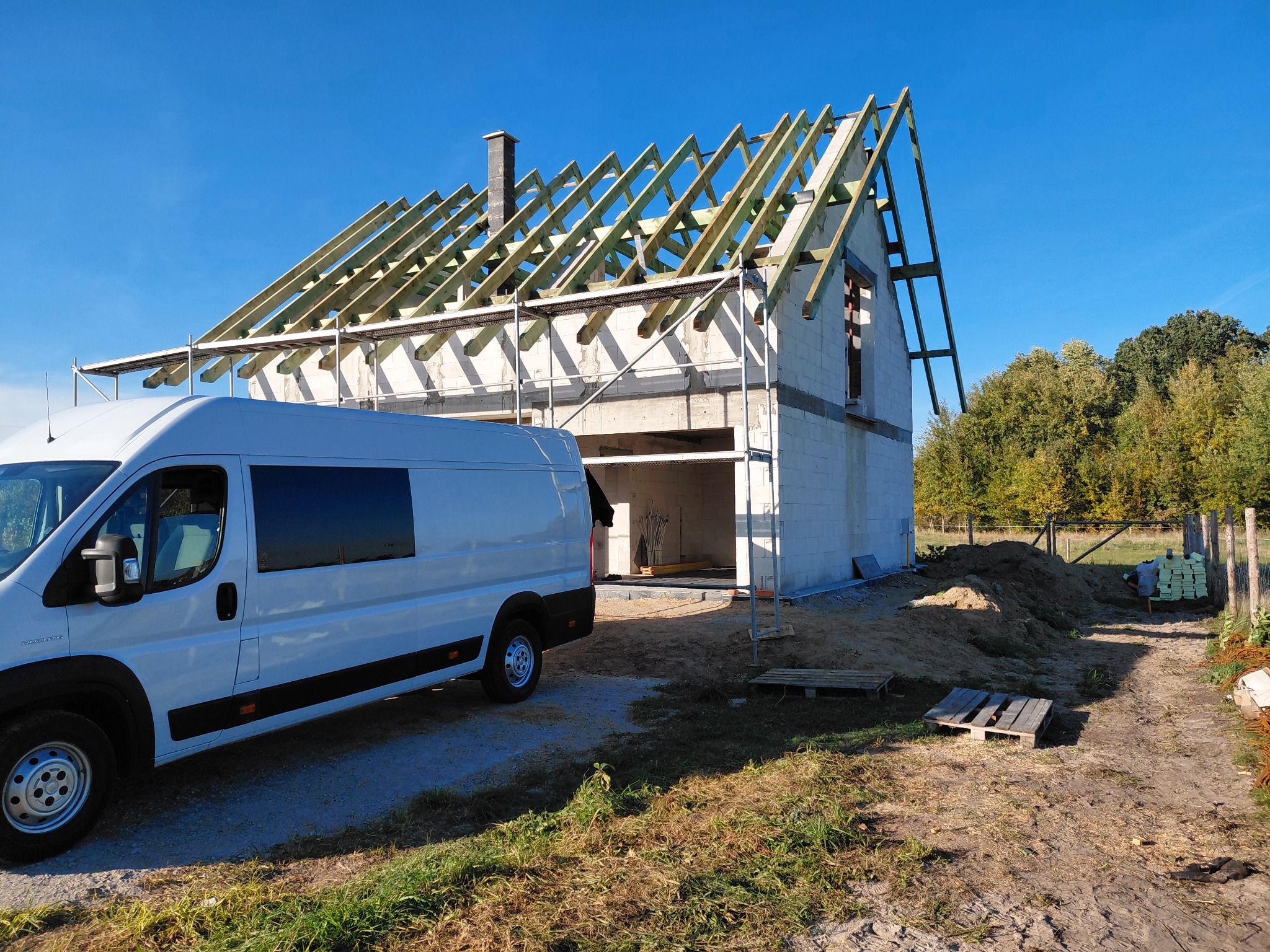 Budowa domu jednorodzinnego w trakcie realizacji: białe ściany z bloczków, drewniana konstrukcja dachu, rusztowania i biały bus dostawczy na tle błękitnego nieba.