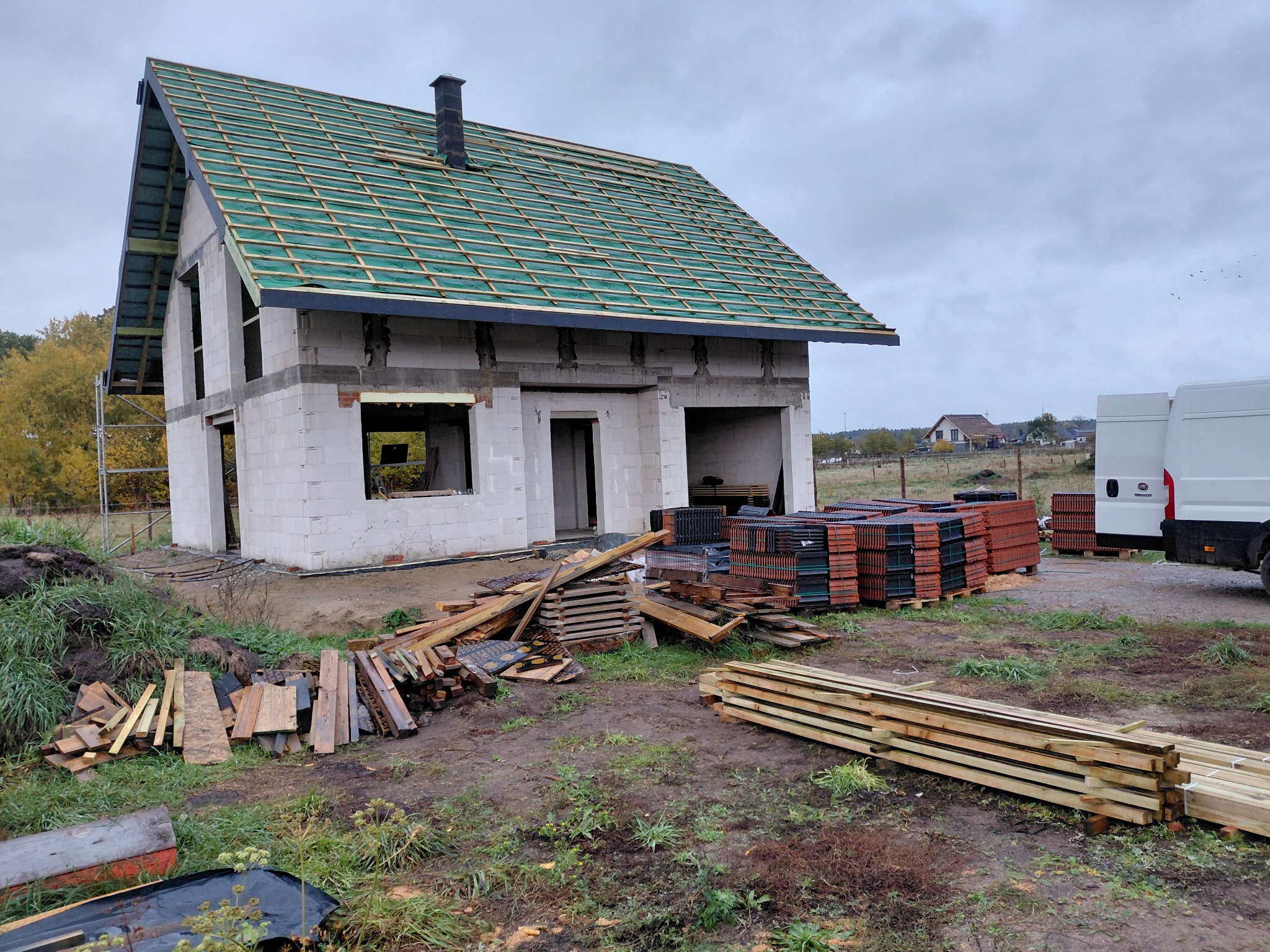 Surowy stan domu z białego pustaka, z zieloną membraną i łatami na dachu, przygotowany do położenia dachówki. Stosy dachówek i drewna budowlanego obok.