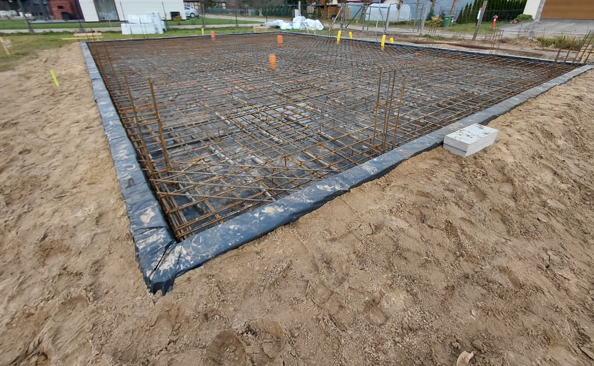 Zbrojenie fundamentu domu jednorodzinnego, widok z góry na siatkę z prętów stalowych, zabezpieczoną folią i obłożoną ziemią. Widoczne pionowe pręty i pomarańczowe rury.