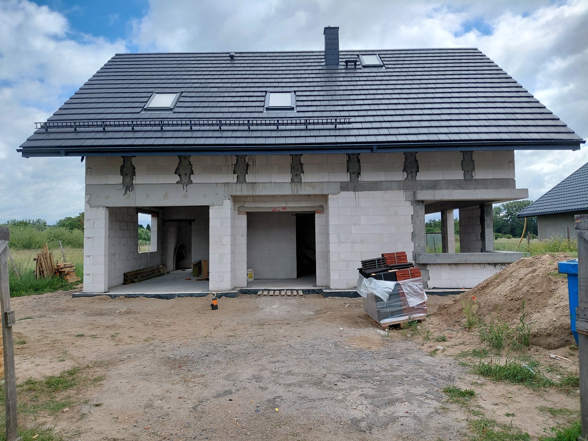 Budynek mieszkalny w stanie surowym otwartym z ciemnoszarym dachem z dachówki, trzema oknami połaciowymi i kominem. Ściany z białych pustaków, widoczne betonowe nadproża, teren budowy z piaskiem...