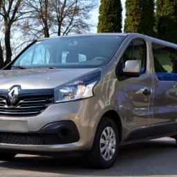 Szary Renault Trafic z niebieskimi siedzeniami pasażerskimi, sfotografowany z zewnątrz na tle drzew.