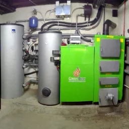 Kompletna kotłownia z dwoma srebrnymi zbiornikami akumulacyjnymi, białym bojlerem, zielonym piecem na paliwo stałe marki GreenHeat i czerwonym zbiornikiem wyrównawczym, liczne rury i zawory...