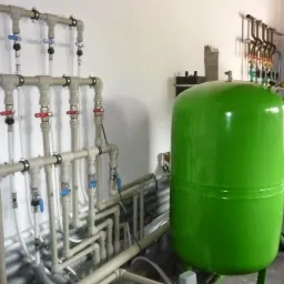 Szczegółowe ujęcie instalacji centralnego ogrzewania z zielonym zbiornikiem wyrównawczym, licznymi rurami i zaworami z czerwonymi i niebieskimi uchwytami, zamontowanej na białej ścianie...