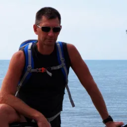 Mężczyzna w okularach i z plecakiem, oparty o rower, z widokiem na morze w tle. Turystyka rowerowa.