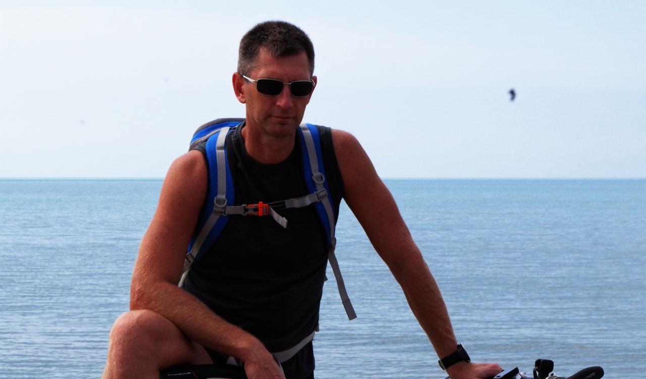 Mężczyzna w okularach i z plecakiem, oparty o rower, z widokiem na morze w tle. Turystyka rowerowa.