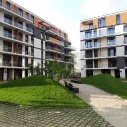 Nowoczesny budynek wielorodzinny z balkonami i elementami drewnianej elewacji, otoczony zielonymi pagórkami i chodnikiem z kostki brukowej.