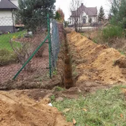 Wąski wykop ziemny wzdłuż ogrodzenia z siatki, świeżo usypana ziemia po obu stronach, widoczne domy w tle, jesienna sceneria z drzewami bez liści.