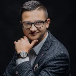 Portret mężczyzny w okularach, w tweedowej marynarce, z zegarkiem na ręce, pozującego na ciemnym tle.