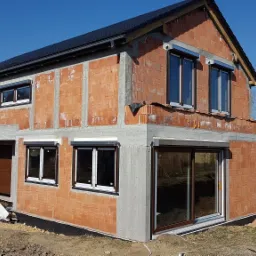 Surowy budynek w trakcie budowy z zamontowanymi oknami z roletami zewnętrznymi, widoczne cegły, betonowe elementy konstrukcyjne i przygotowane podłoże.