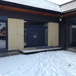 Nowoczesny dom jednorodzinny z płaskim dachem, antracytową elewacją, jasnymi panelami dekoracyjnymi, dużymi oknami z roletami zewnętrznymi i dwuskrzydłowymi drzwiami wejściowymi z metalu w zimowej...