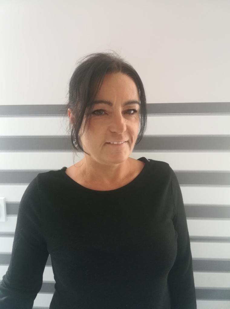 Portret kobiety w czarnej bluzce na tle ściany w poziome pasy, uśmiechnięta, z ciemnymi włosami i delikatnym makijażem.