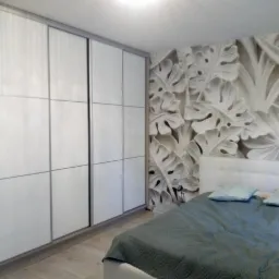 Szafa przesuwna z białymi panelami w sypialni z dekoracyjną tapetą w liście monstery, widoczny fragment łóżka i dziecięcego łóżeczka.