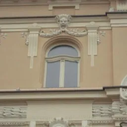 Elewacja budynku z oknem w stylu klasycystycznym, jasnobeżowa fasada z dekoracyjnymi sztukateriami, okno łukowe z białymi szprosami.