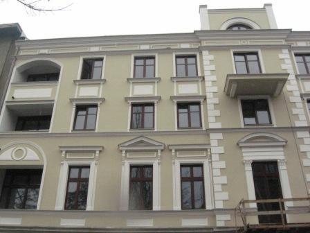 Fasada odnowionego, kilkupiętrowego budynku w stylu kamienicy, z beżową elewacją, białymi zdobieniami wokół okien i balkonów, widoczne okna z ciemnymi ramami.