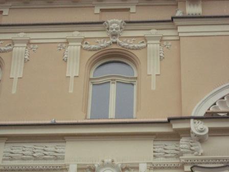 Elewacja budynku z oknem w stylu klasycystycznym, jasnobeżowa fasada z dekoracyjnymi sztukateriami, okno łukowe z białymi szprosami.