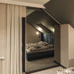 RAVATTI - Nowoczesna sypialnia na poddaszu z łóżkiem z welurowym zagłówkiem, widoczna przez szerokie drzwi z zasłonami, z fragmentem drugiego łóżka na pierwszym planie.