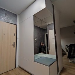 RAVATTI - Biała zabudowa w przedpokoju z siedziskiem z szarymi poduszkami i lustrem na całej ścianie, odbijającym osobę robiącą zdjęcie telefonem.