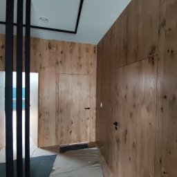 RAVATTI - Korytarz wykończony drewnianą boazerią z ukrytymi drzwiami, czarne, minimalistyczne poręcze i listwy sufitowe, podłoga zabezpieczona folią malarską.