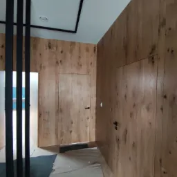 Korytarz wykończony drewnianą boazerią z ukrytymi drzwiami, czarne, minimalistyczne poręcze i listwy sufitowe, podłoga zabezpieczona folią malarską.