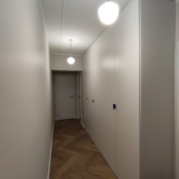 RAVATTI - Wąski korytarz z zabudową szaf wnękowych o minimalistycznym designie, drewnianą podłogą ułożoną w jodełkę i dwoma okrągłymi lampami sufitowymi.