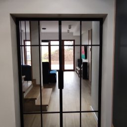 RAVATTI - Szklane drzwi przesuwne z czarnymi szprosami dzielące przestrzeń, widok na schody i salon z drewnianą podłogą.