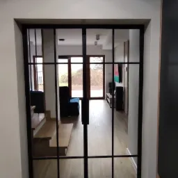 Szklane drzwi przesuwne z czarnymi szprosami dzielące przestrzeń, widok na schody i salon z drewnianą podłogą.