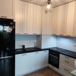 RAVATTI - Nowoczesna kuchnia z białymi szafkami, czarnym blatem i parkietem ułożonym w jodełkę. Oświetlenie sufitowe z geometrycznym kloszem tworzy promieniujący wzór cieni na suficie.