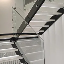 Nowoczesne schody z białymi stopniami i czarną konstrukcją, zabezpieczone szklaną balustradą mocowaną na metalowych uchwytach.