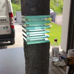 RAVATTI - Dekoracyjna kolumna z szarego kamienia z wstawkami z przezroczystego, zielonkawego szkła, na stole warsztatowym z narzędziami i butelką płynu do czyszczenia, z widocznym fragmentem samochodu...