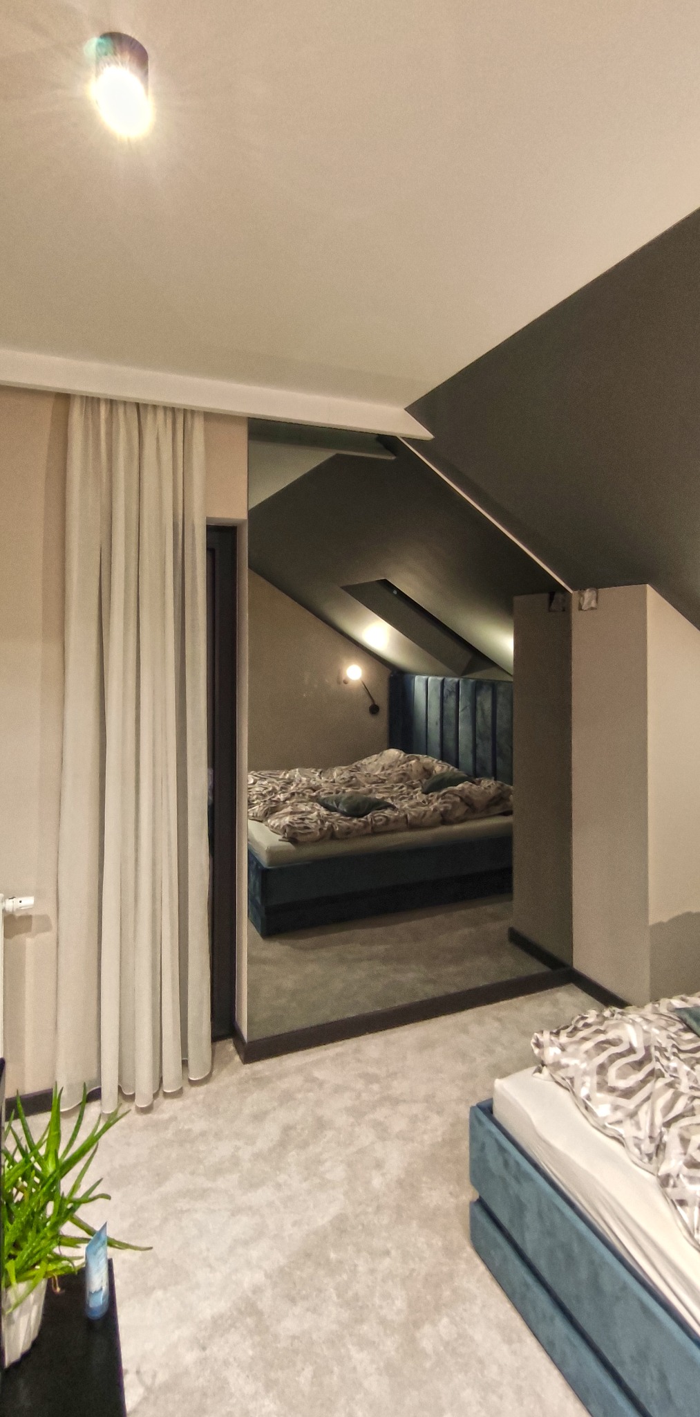 Nowoczesna sypialnia na poddaszu z łóżkiem z welurowym zagłówkiem, widoczna przez szerokie drzwi z zasłonami, z fragmentem drugiego łóżka na pierwszym planie.