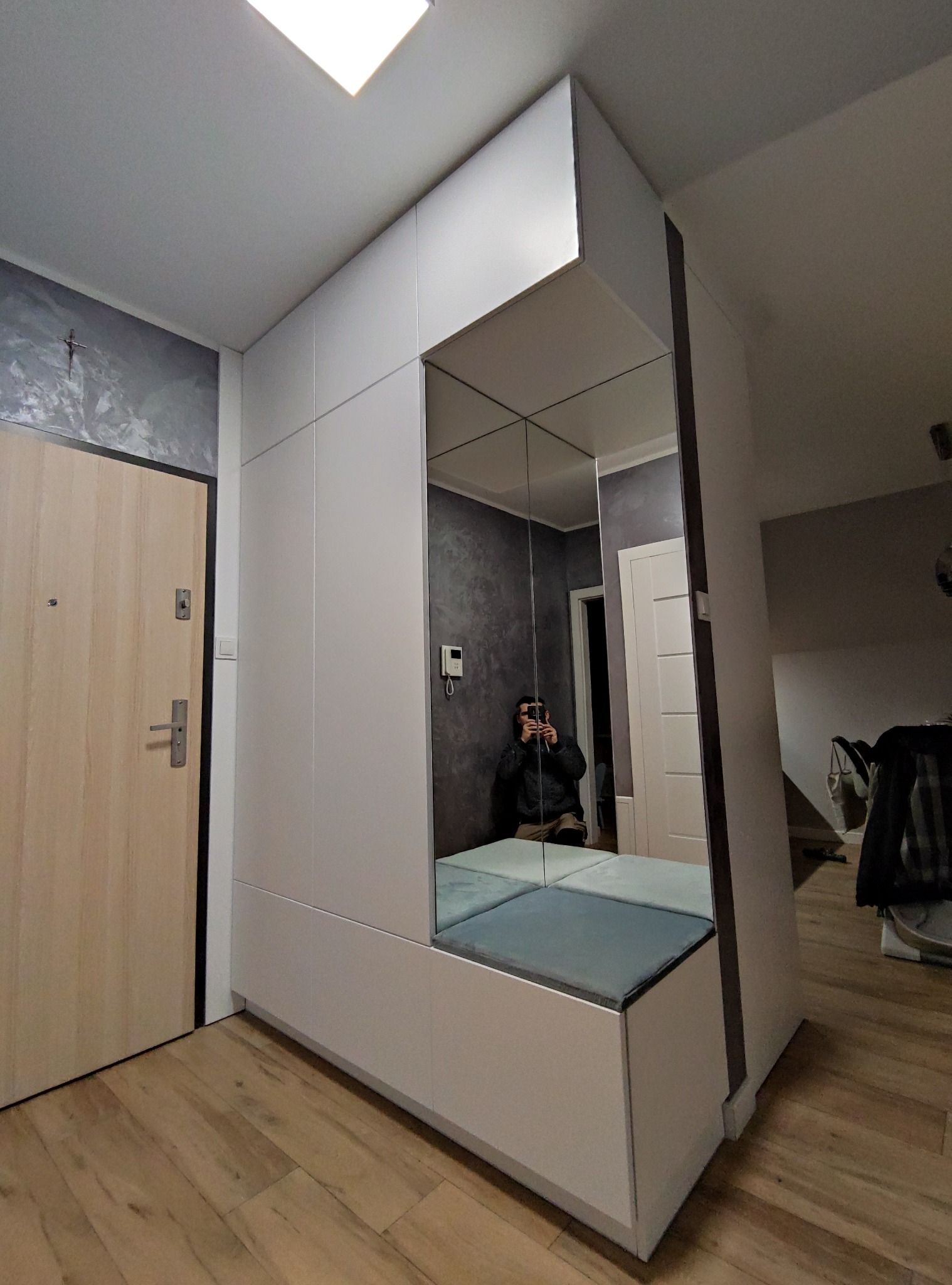 Biała zabudowa w przedpokoju z siedziskiem z szarymi poduszkami i lustrem na całej ścianie, odbijającym osobę robiącą zdjęcie telefonem.