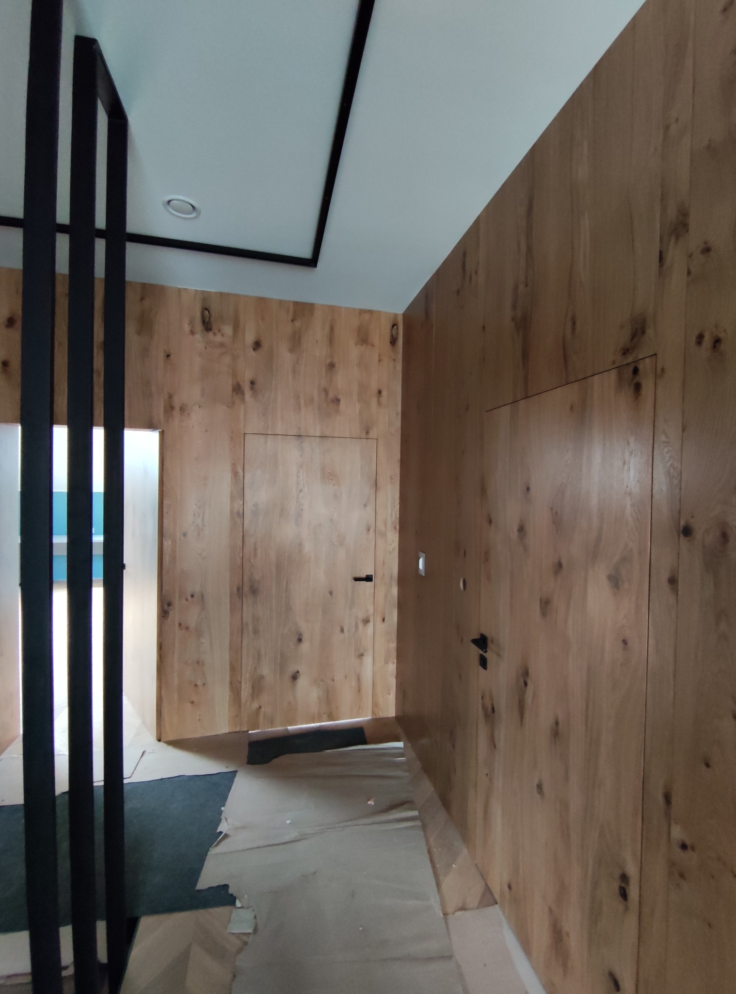 Korytarz wykończony drewnianą boazerią z ukrytymi drzwiami, czarne, minimalistyczne poręcze i listwy sufitowe, podłoga zabezpieczona folią malarską.