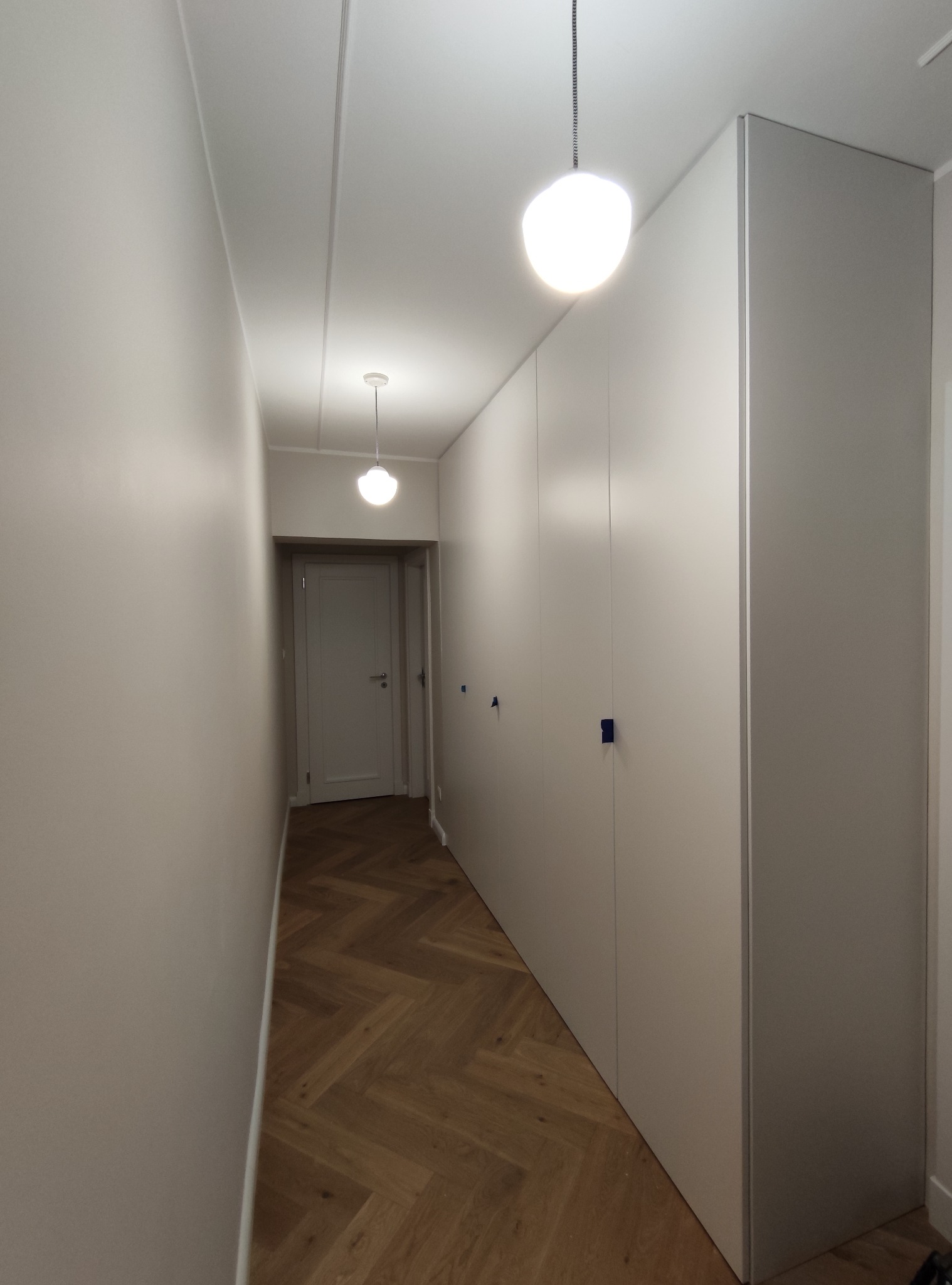 Wąski korytarz z zabudową szaf wnękowych o minimalistycznym designie, drewnianą podłogą ułożoną w jodełkę i dwoma okrągłymi lampami sufitowymi.