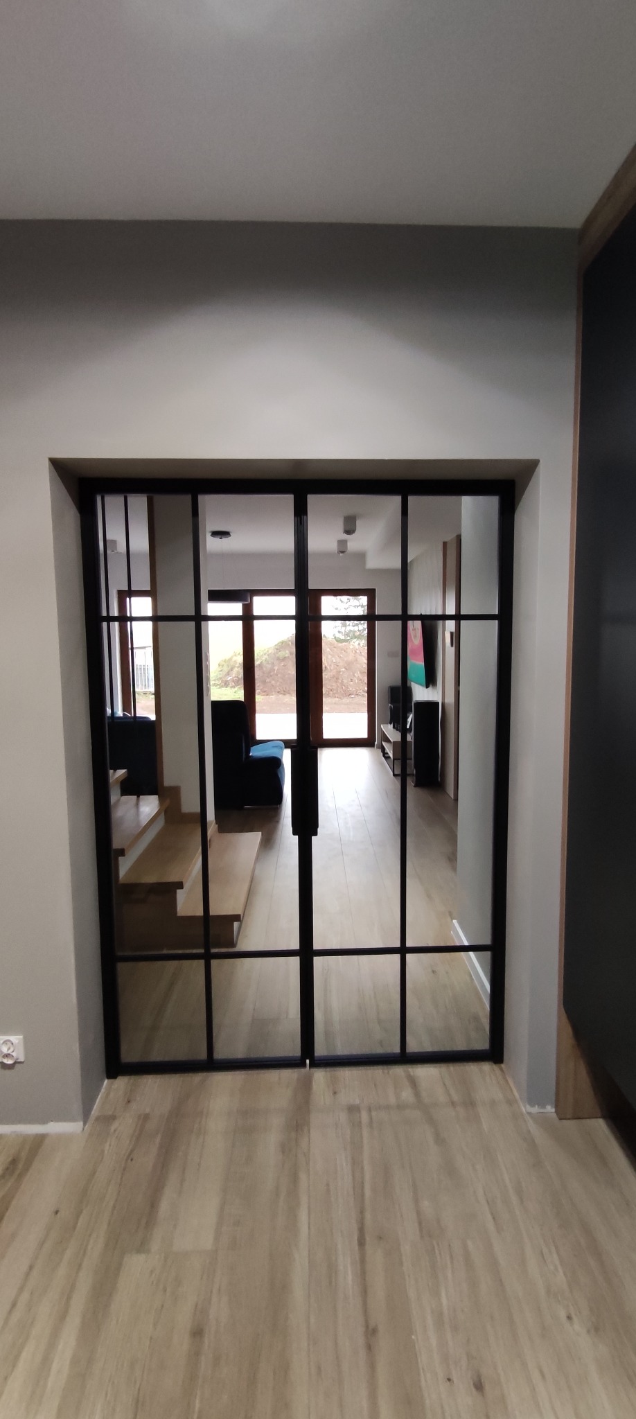 Szklane drzwi przesuwne z czarnymi szprosami dzielące przestrzeń, widok na schody i salon z drewnianą podłogą.