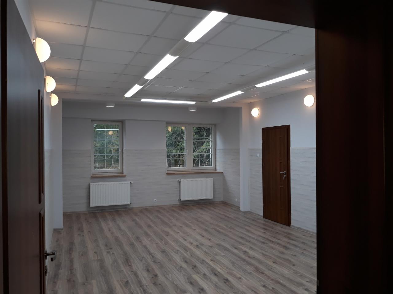 Odnowione wnętrze biura w Białymstoku, widok na świeżo pomalowane ściany w odcieniach szarości, drewnopodobną podłogę, grzejniki pod oknami i nowoczesne oświetlenie sufitowe.