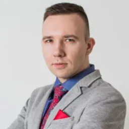 Portret mężczyzny w szarej marynarce, błękitnej koszuli i wzorzystym krawacie z czerwoną poszetką. Jasne tło, stonowane światło, skupienie na twarzy i wyrazie.